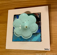 Front view showing white square frame, blue collage background, mint green ceramic flower, signature "mau"