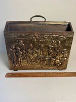 Front view showing embossed brass panel scene and ruler for scale
