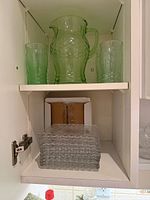 Green glass pitcher, two tumblers, stack of clear square plates in cabinet