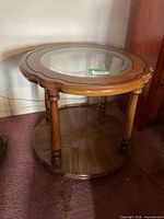Three-quarter view of round wooden end table showing glass top, turned legs, and lower shelf
