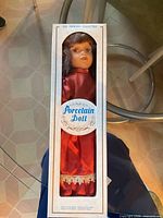 Front view of boxed porcelain doll showing red dress and branding