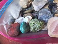 Assorted stones showing red jasper, green chrysocolla/malachite, peridot rough, polished pink stone and other mixed rocks in plastic bag
