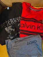 Three T-shirts laid flat: black Aftershock, red Calvin Klein, grey PlayStation