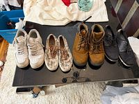 Five pairs on table – white leather sneakers, floral canvas, brown boots, black strap sneakers, black dress shoes