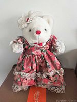 Front view of white plush teddy bear in plaid floral dress and bonnet standing on metal stand