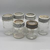 Front view of five jars showing differing heights, embossed fronts, and metal lids