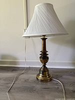 One brass table lamp with pleated white shade resting on floor, full view including cord