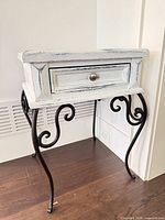 Front and side view of distressed white wood side table showing drawer and wrought iron scroll legs