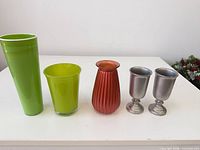 Front view of all five decorative pieces: two green glass vases, one red ribbed glass vase, two silver metal goblets