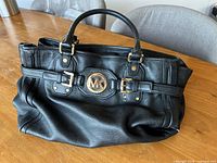 Front view showing black leather, MK logo medallion, buckle details, handles