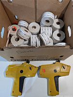 Box containing multiple white label rolls beside two yellow Monarch 1110 price guns