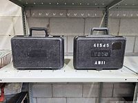 Front view of both black hard plastic cases on shelf showing handles, latches, stenciled ID on left case