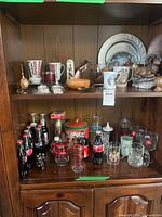 Overall view showing Coca-Cola bottles, tins and decorative items on lower shelf and mugs, figurines on upper shelf