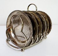 Seven etched silver-tone coasters standing in wire holder, full view