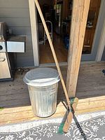 Push broom leaning against post, garbage can with lid visible