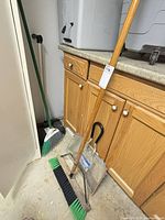 Wide wooden push broom, angled plastic broom, metal dustpan visible beside cabinetry
