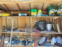 Overview of three shelves with herbicide jugs, fertilizer tub, planter, pail, shovel, saw, fittings, clock