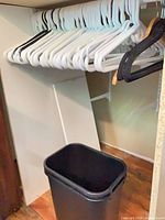 White and black plastic hangers hanging on rod with black wastebasket below