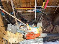 Assorted tools on shed floor: brooms, shovels, orange leaf blower, Yardworks handles and attachments