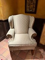Front view of beige patterned wingback chair showing upholstery, rolled arms, nailhead trim, tapered legs