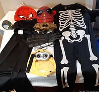 All costume items laid out on table including skeleton jumpsuit, Spider-Man masks, Minion hat, Batman cape, mask and emblem, gloves