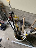 Bucket containing assorted long-handled tools with shovel and ladders nearby