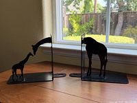 Two black metal bookends showing giraffe silhouettes on wooden surface by window