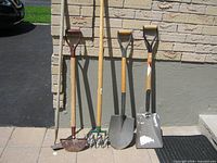 Five long-handled garden tools leaning against wall