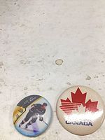 Front view of both pinback buttons showing Canada maple leaf design and hockey player design