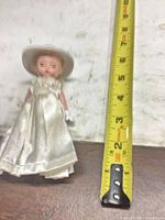 Front view of doll beside ruler showing approx 4 in height, ivory dress, hat, gloves