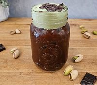 Iced beverage in mason jar topped with chopped chocolate pieces, pistachios scattered on table