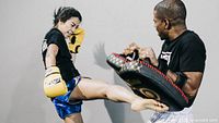 Participants practicing Muay-Thai kick with trainer holding pads – studio branding visible