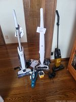 Group of three stick vacuums, one handheld vacuum, chargers and accessories on floor