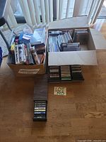 Overall view: two cardboard boxes with CDs and VHS tapes, cassette drawer racks on floor