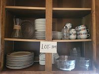 Full cabinet view showing stacked porcelain plates, bowls, cups, Chemex-style carafe, monogrammed glass bowl, ramekins and glasses