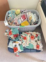 Laundry basket and additional stack showing assortment of kitchen linens with various patterns and colors