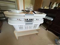 Two stacked white laundry baskets filled with assorted hangers sitting on small white shelf