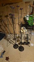 Group view of multiple metal stands including three dark bronze floor stands, two wire frames, brass candlestick and black pot holder