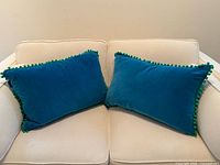 Two blue velvet pillows with green pom pom trim on cream sofa
