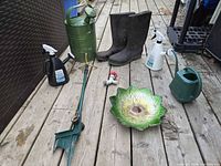 All lot items arranged on deck: metal and plastic watering cans, rubber boots, sprinkler spike, spray bottles, glass bird bath bowl, faucet valve