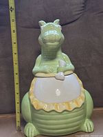 Front view of green ceramic dinosaur cookie jar with measuring tape showing height