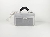 Closed silver Cosmepak hard-shell makeup train case showing front ribbed panel, latch, handle, and clear strap