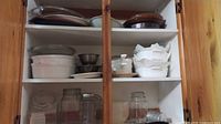 Overview of cabinet shelves showing CorningWare dishes, glass bakeware, mixing bowls, storage jars and hand tool