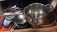 Cookware items in cabinet including saucepan, pot, lids, steamer insert, colander