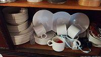 Shelf view showing stacks of white plates, saucers, cups, triple-well platter and additional items