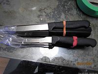 Three of the knives stacked, showing stainless blades, black handles, rubber bands and plastic sleeve