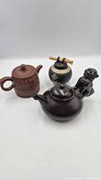 Three teapots grouped: brown clay, dark brown with lion, black ceramic with bamboo handle
