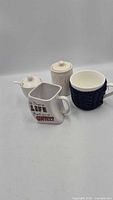 Group shot of four ceramic items: square printed mug, round mug with navy sleeve, small lidded teapot/creamer, knit-texture lidded jar