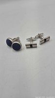 Displays both pairs of cufflinks: round blue inlay pair and rectangular black-striped pair