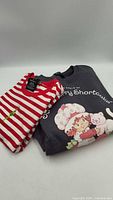 Folded Strawberry Shortcake sweatshirt beside folded red-white striped Grinch t-shirt